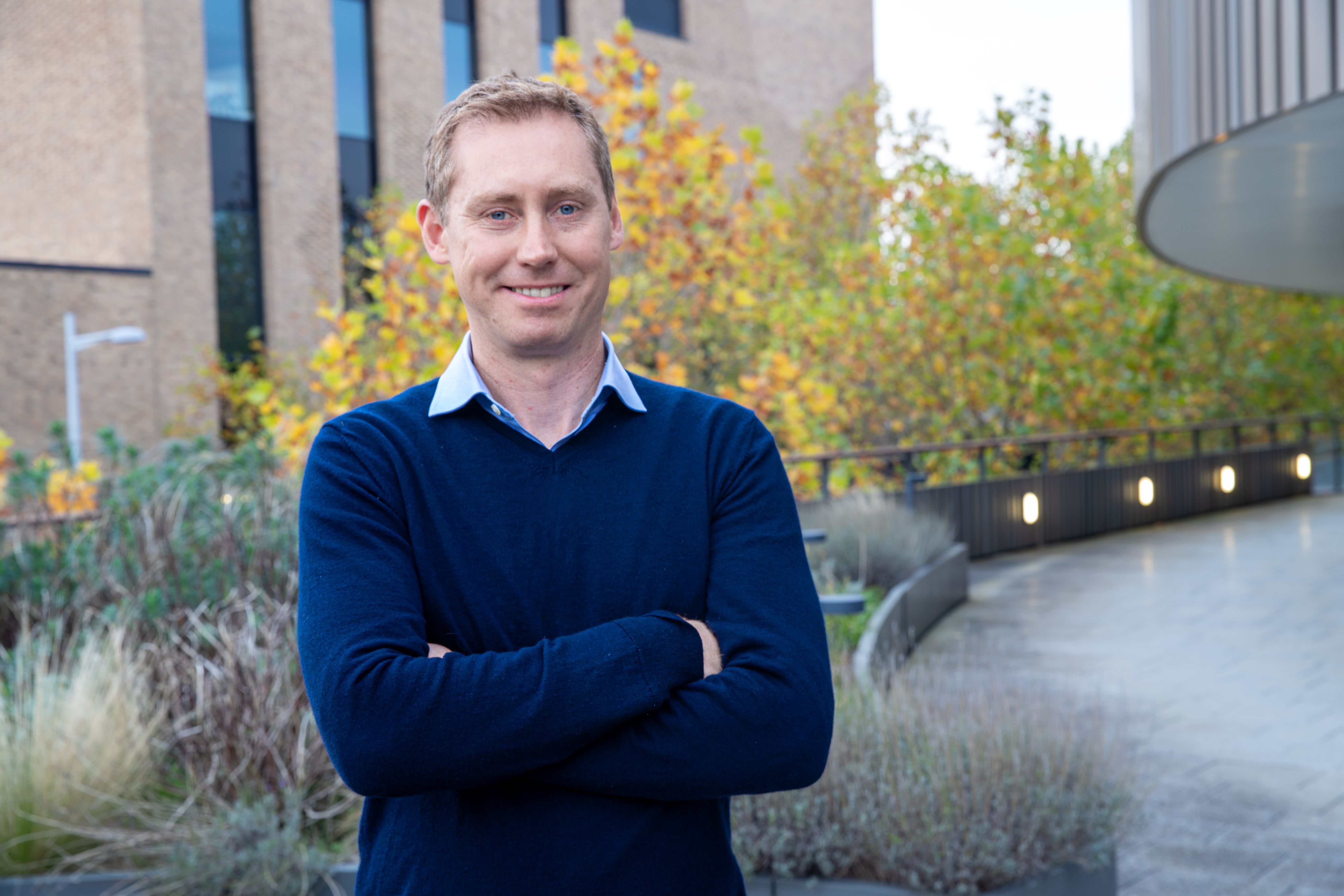 Sam Stranks with crossed arms smiling at the camera in a blue jumper and shirt, outside CEB with green plant beds behind.