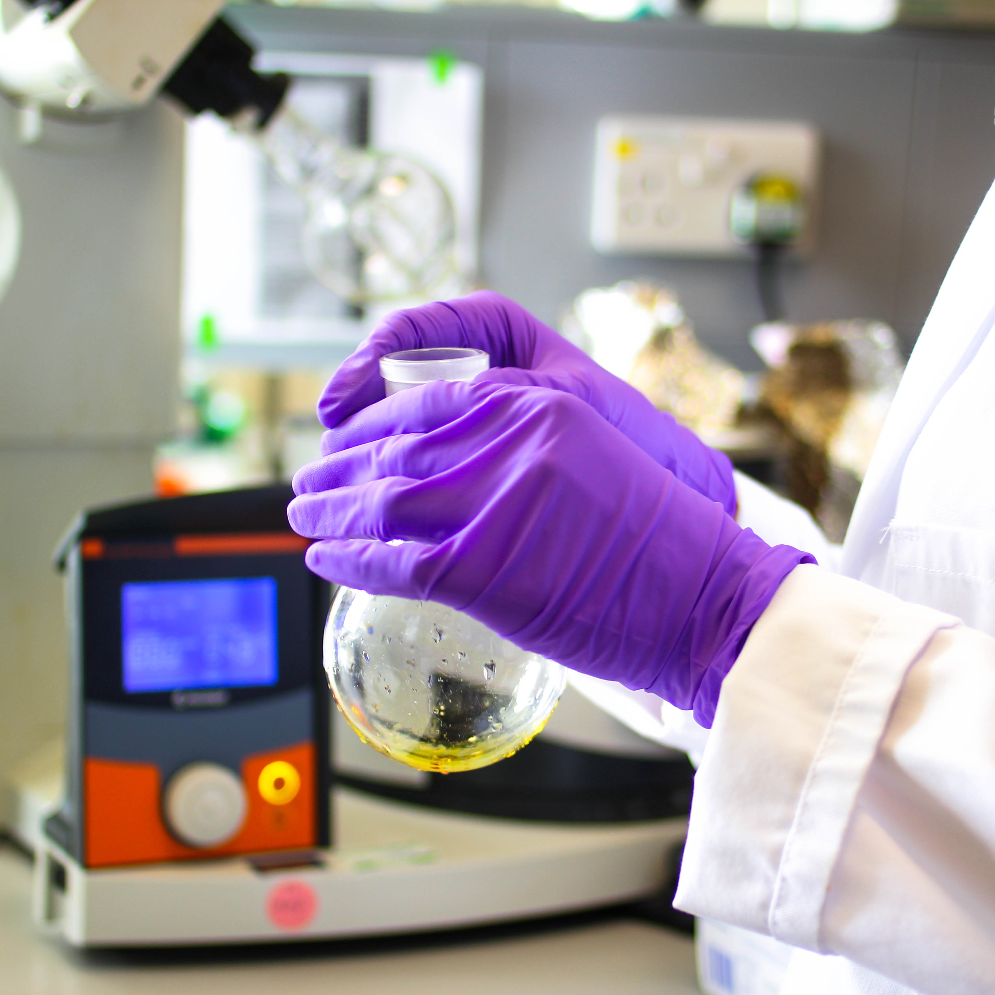 Purple gloved hands of person in white lab coat holding test tube with yellow liquid in bottom.