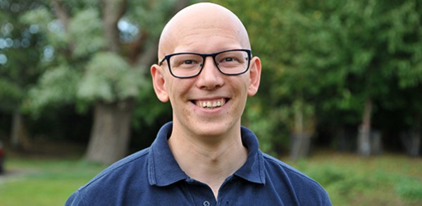 Bald man with glasses smiling into camera, head and shoulders shot. Trees out of focus in background