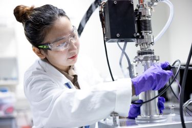 Woman with safety glasses, white lab coat and purple plastic gloves working with lab equipment/machine