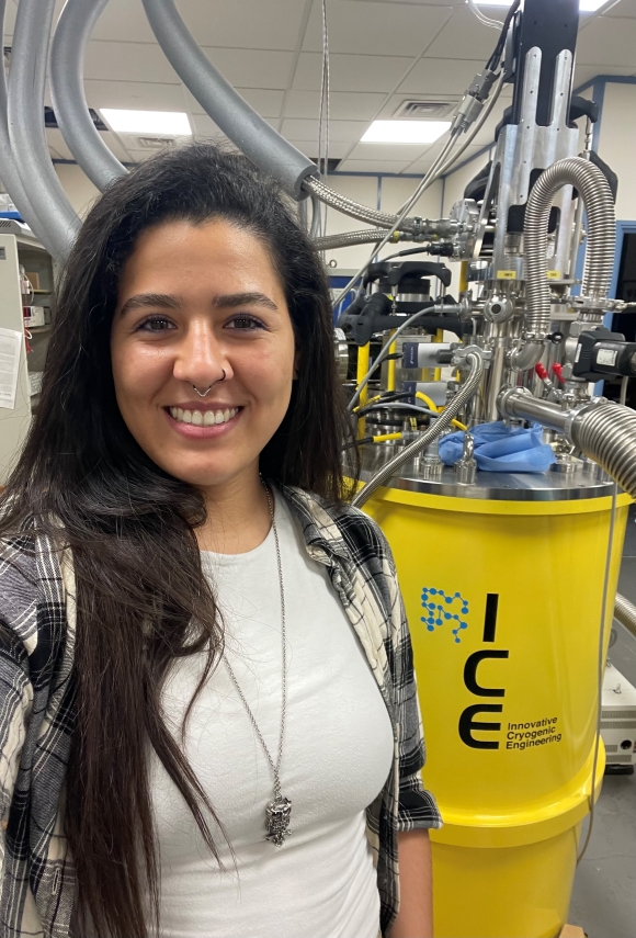 Woman smiling at the camera with long dark hair, with a bright yellow barrel in background with ICE on it