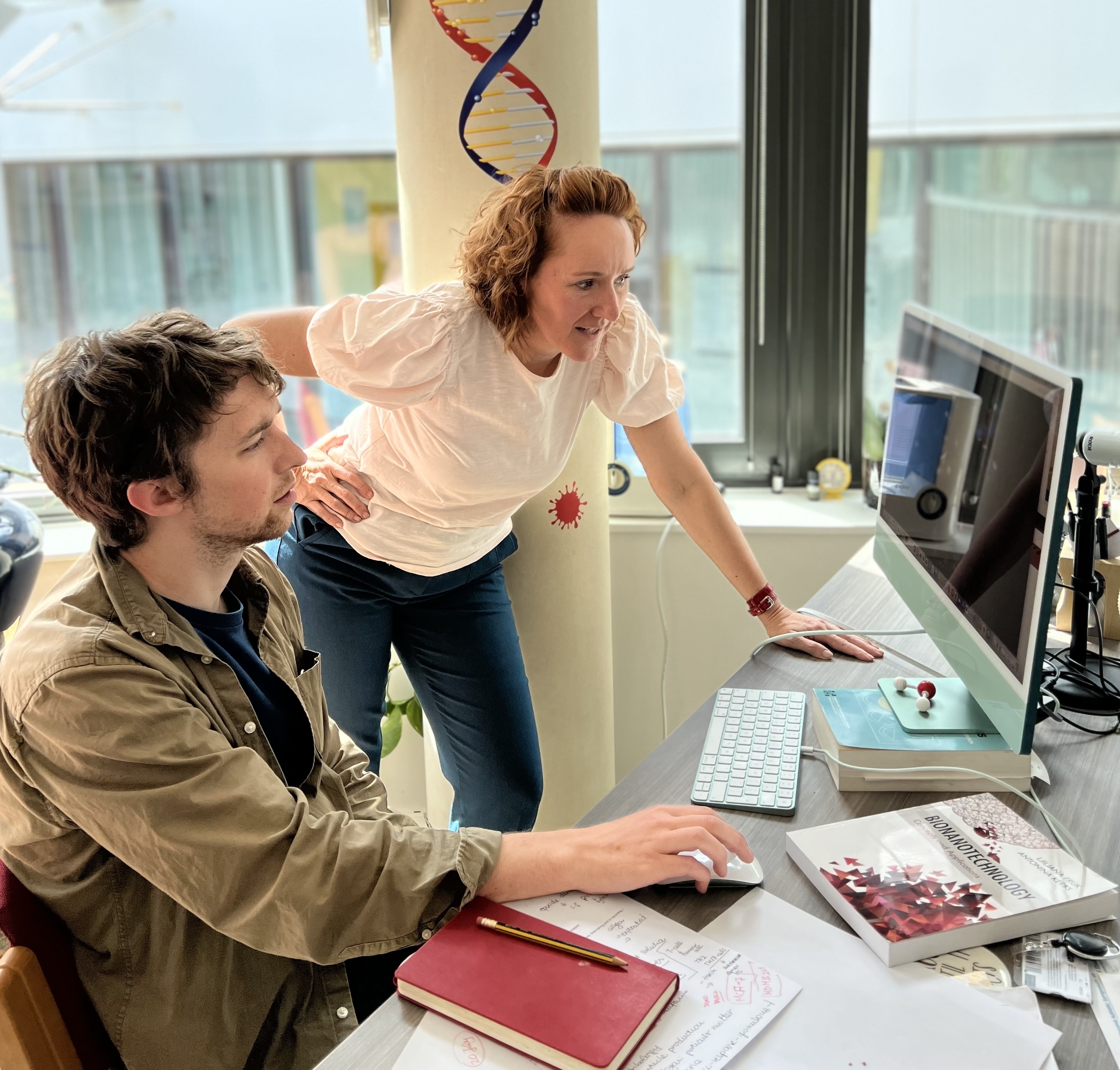 Professor LKjiljana Fruk stood with one hand on hip while leaning on desk looking at computer screen with Andrew sat down with hand on mouse