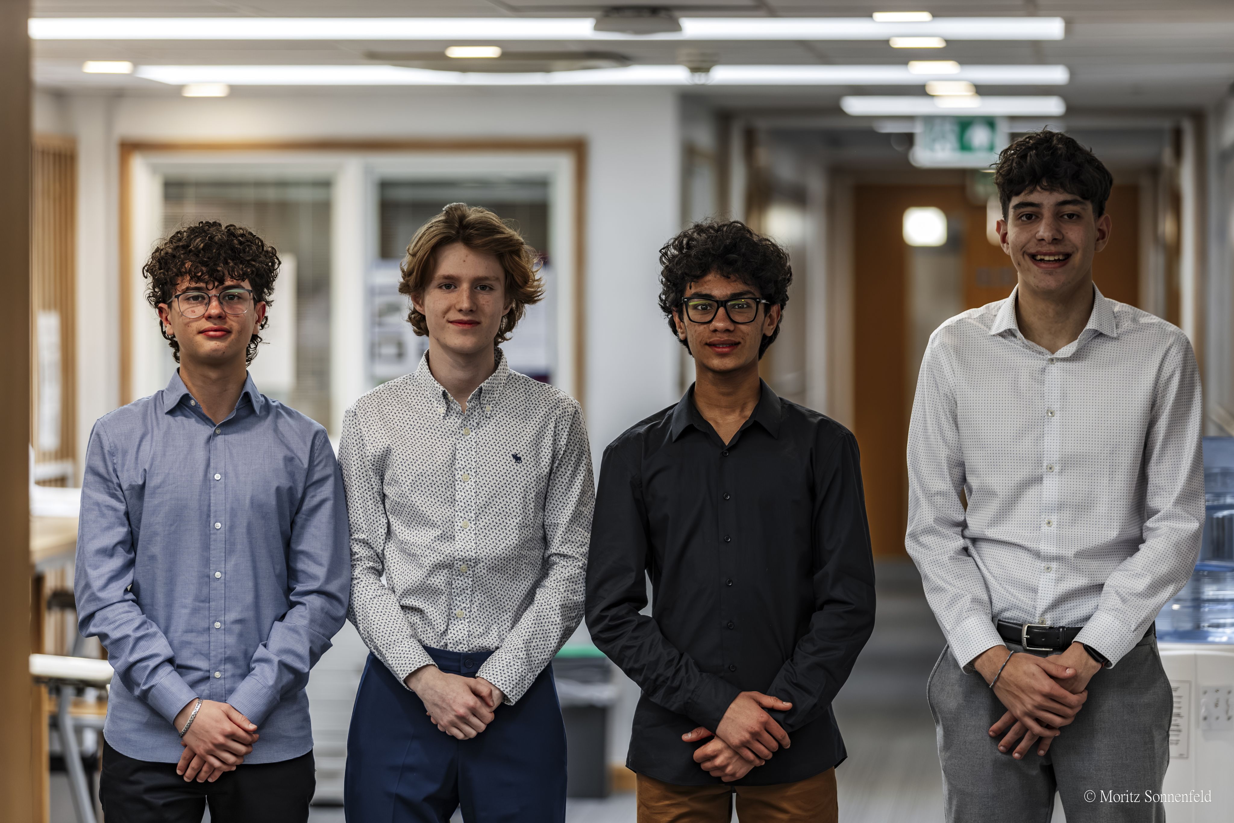 Four young men in shirts in an office/school setting stood formally