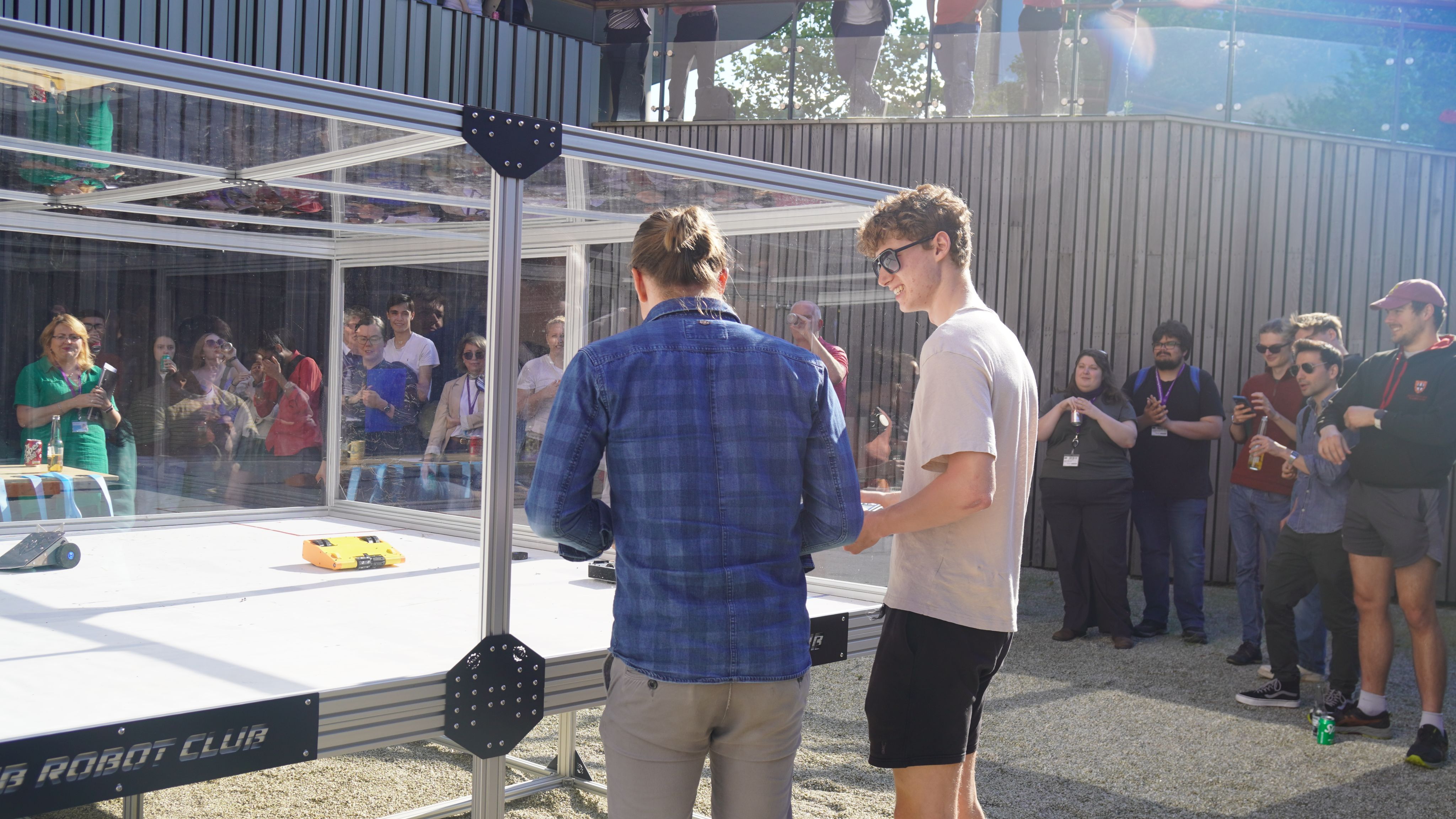 Two men in casual clothing outside in the sun with a large glass fronted robot arena in front, and people visible round the edge of the space.