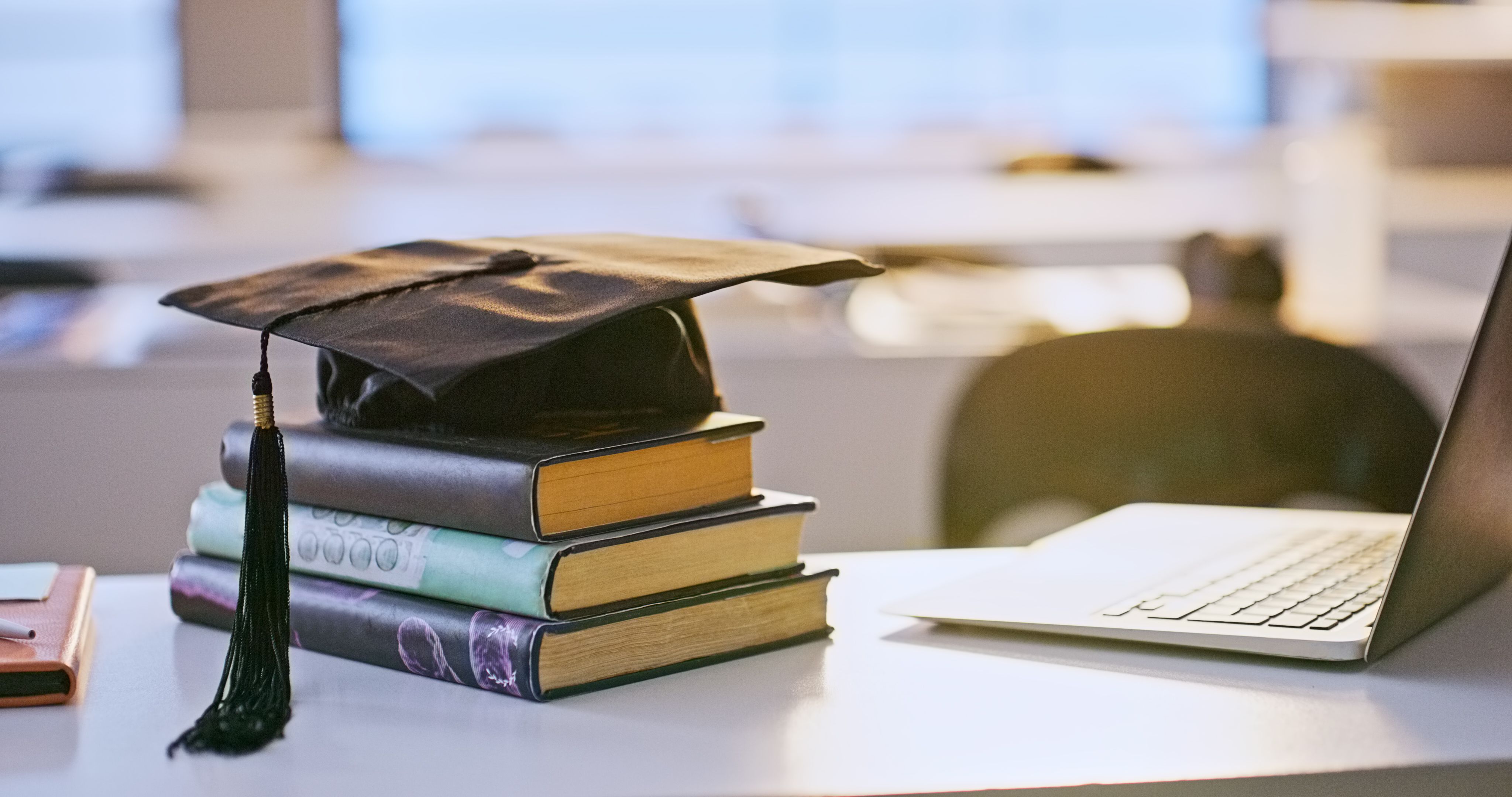 Three hardback books stacked on a desk with a graduate mortarboard on top, a silver laptop sits on its left.