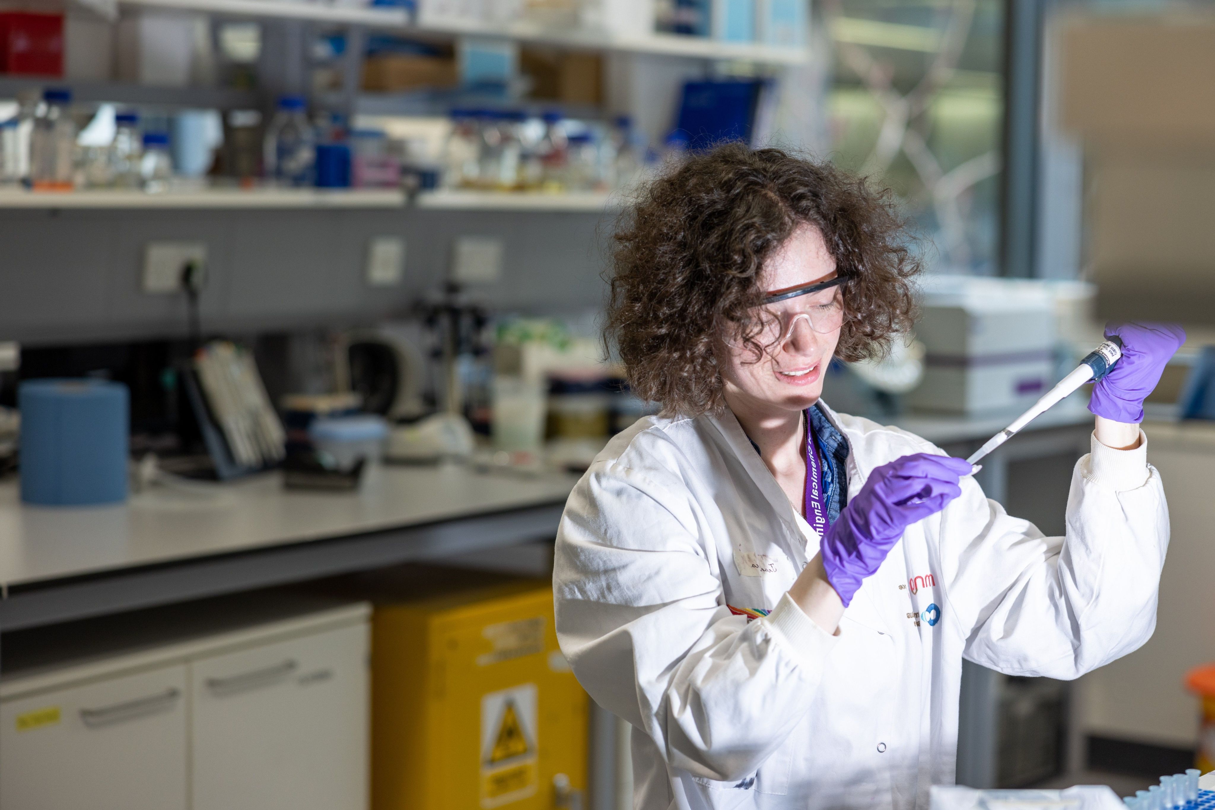 Woman in white lab coat, goggles and gloves pipettes in a lab environment.