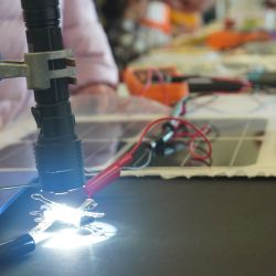 Torch shining brightly on a small pinned solar square connected to red wires that disappear into background