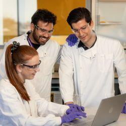 One woman (seated) and two men, in white labs, look relaxed and friendly as they look at a laptop in a lab setting.
