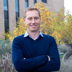 Sam Stranks with crossed arms smiling at the camera in a blue jumper and shirt, outside CEB with green plant beds behind.