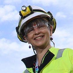 Fiona Macleod looking down at the camera with a blue sky with clouds, dressed in high vis and a hard hat with torch attached.