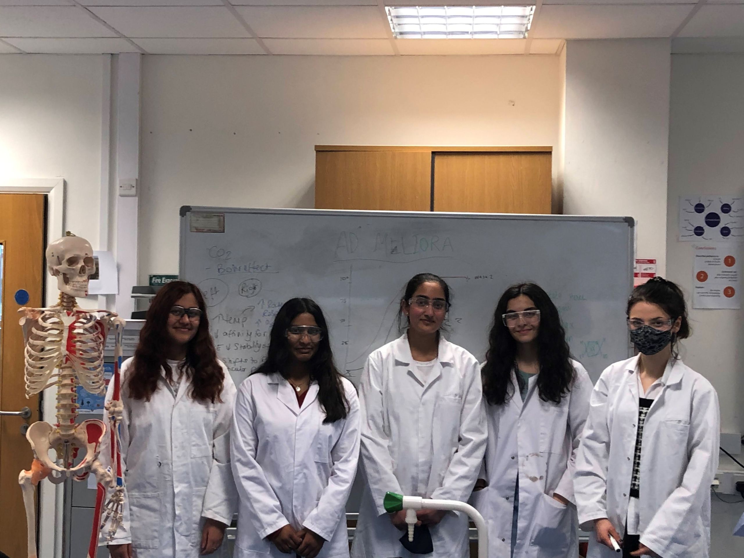 Five female students in lab coats stand in front of a whiteboard in their science classroom