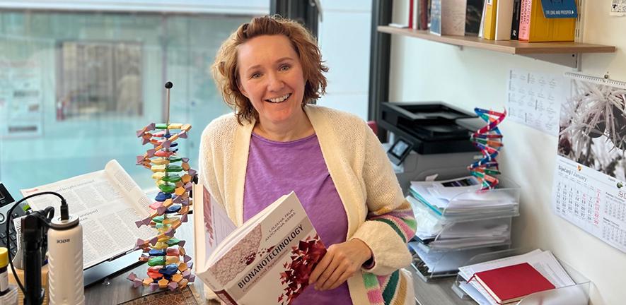 Ljiljana Fruk in her office holding the Bionanotechnology textbook and standing next to a model of DNA