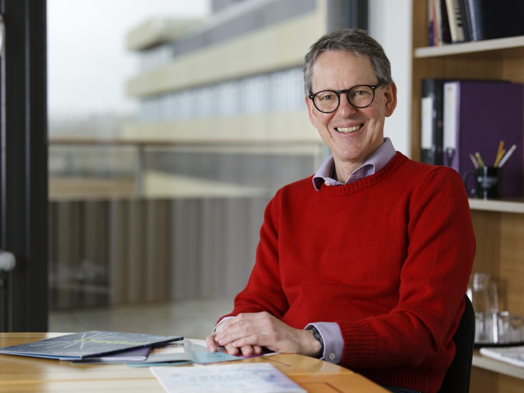 A man in a red top sits at a table and smiles towards the camera.