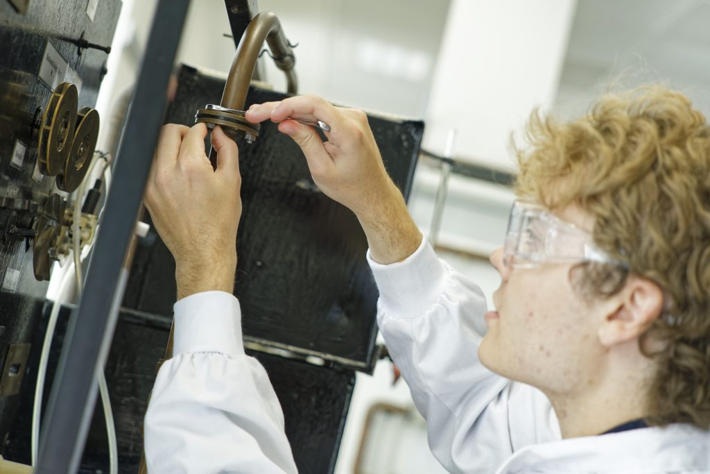 Man in white lab coat and safety goggles fixing mechanical engineering equipment in lab setting.