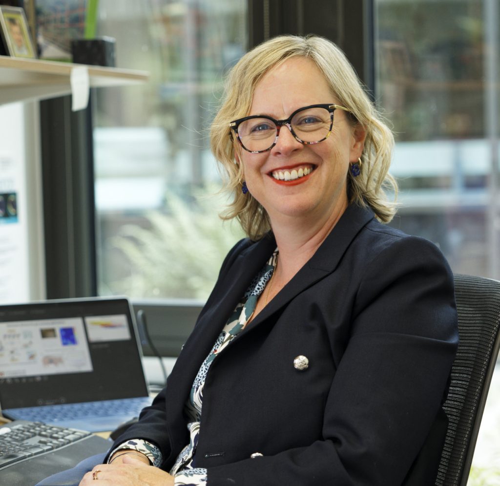A blonde women sitting in a modern office, smiling at the camera.