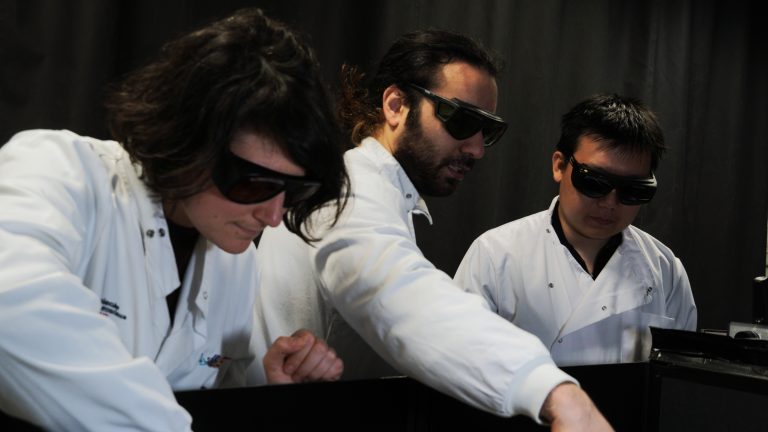 Three people in lab coats and laser goggles peer down into a box.