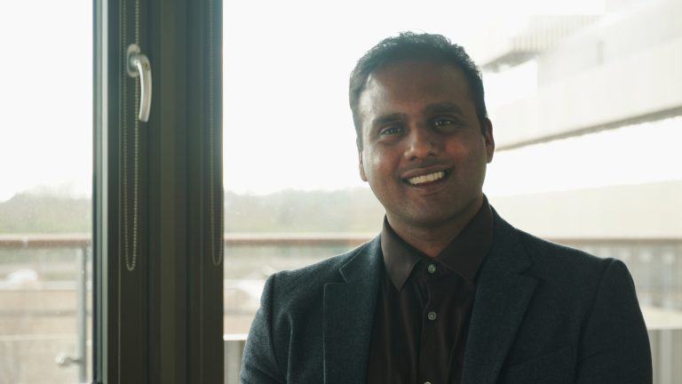 Akshay Deshmukh smiles at the camera in dark suit with West Cambridge in background.