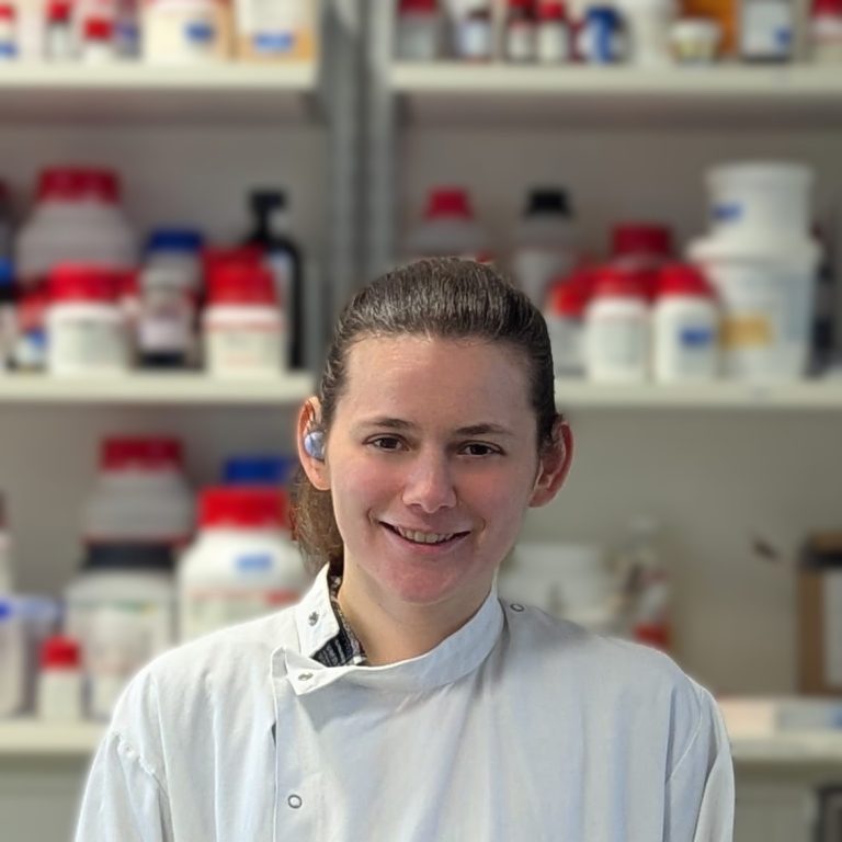 Portrait of a woman in a lab coat smiling at the camera. A lab environment forms the background.