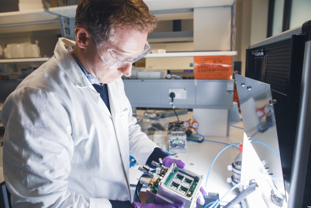 Sam Stranks in white lab coat and purple gloves looks at a piece of equipment in the lab, illuminated with light from a computer on right of picture.