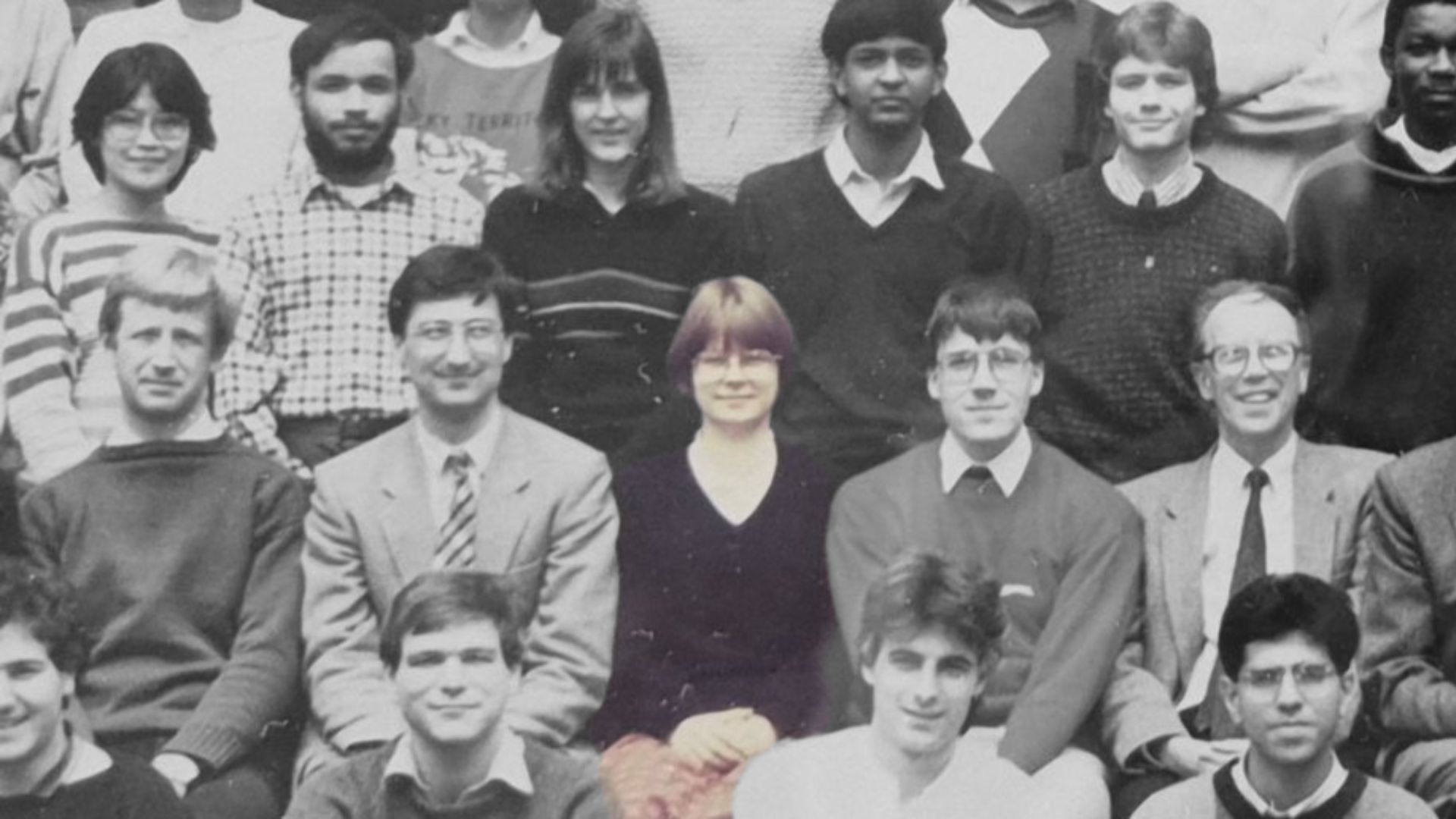 A black and white image showing multiple rows of people facing the camera. One young woman is highlighted in colour.