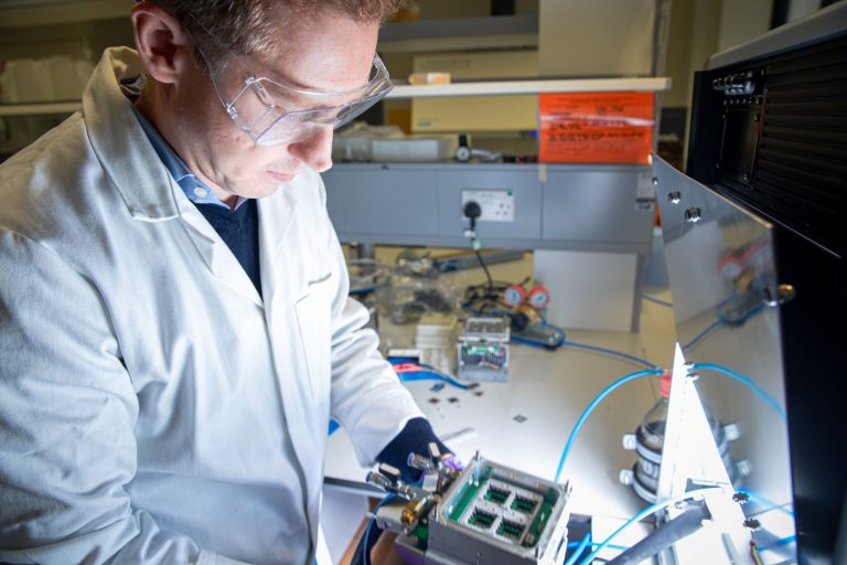 Close up shot of Sam Stranks at work in the lab, in a white lab coat and holding a piece of equipment.