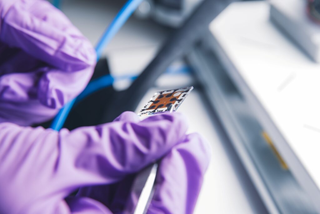 Purple gloved hand holds small chip like device, a perovskite, with wires and lighting in background.
