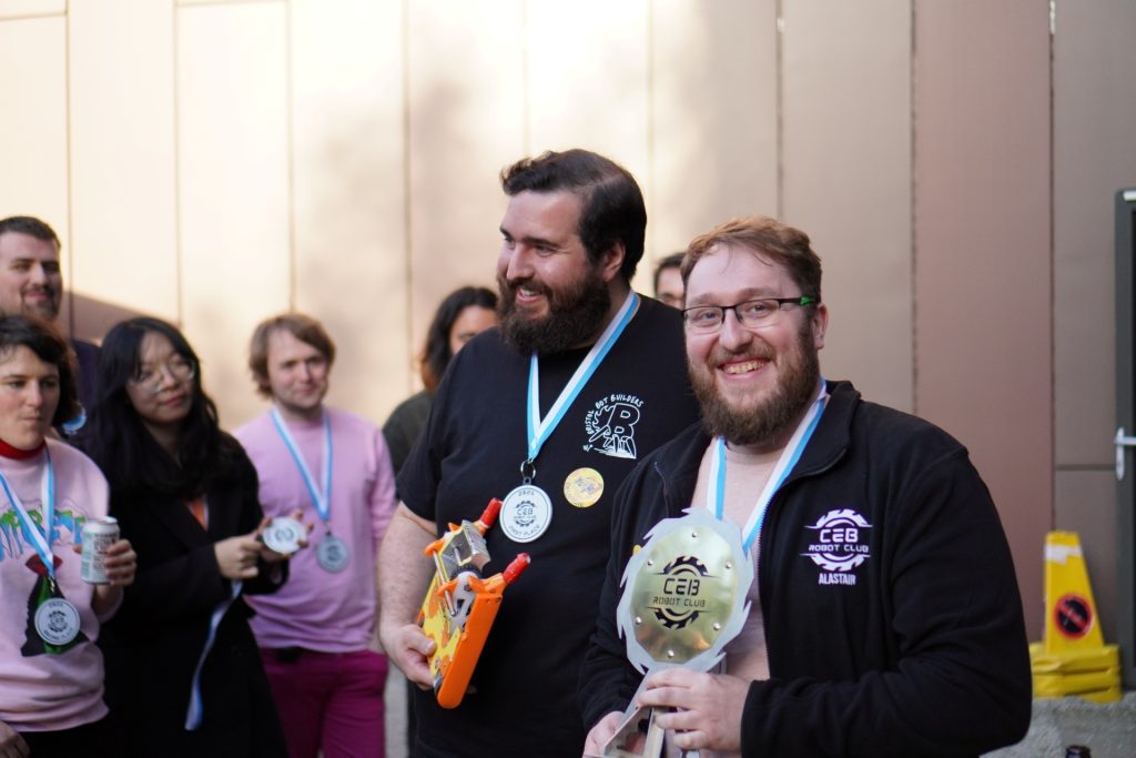 Lewis Reeves and Alastair Robinson smile with their winning trophy and robot, outside, with people in background.