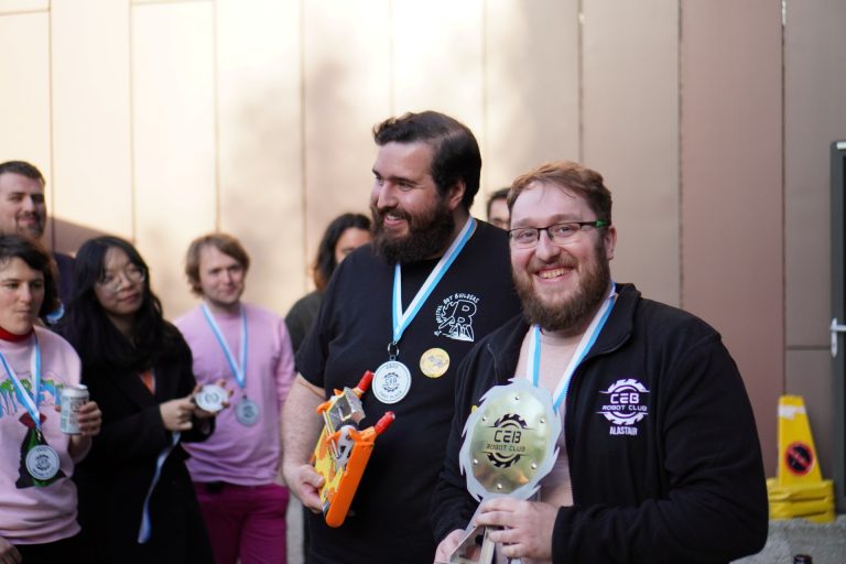 Lewis Reeves and Alastair Robinson smile with their winning trophy and robot, outside, with people in background.