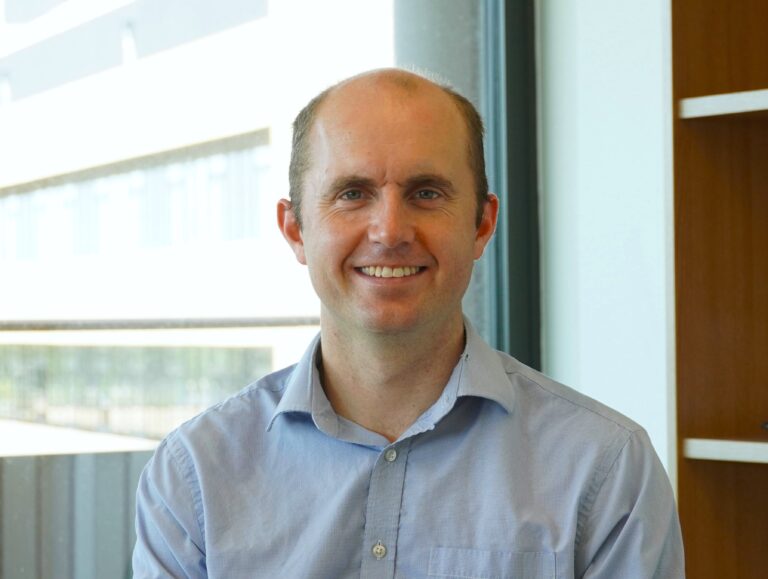 Dr George Gordon in light blue, open button shirt smiling at the camera in office setting.