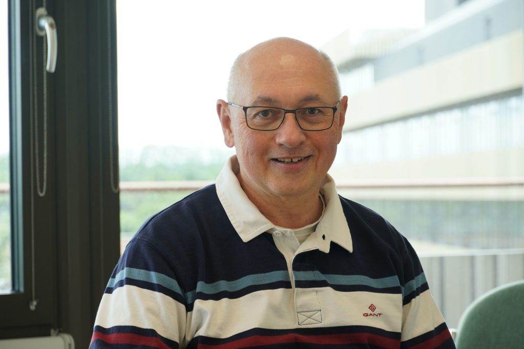 Markus Kraft headshot in CEB office with MSM in the background. In blue striped polo shirt.